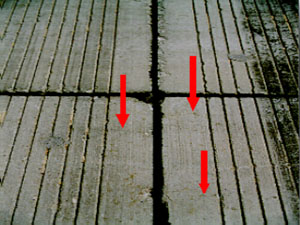 Close-up photo showing multiple small hairline shrinkage cracks on the pavement surface.            Three red arrows are drawn on the photo to point to the location of the visible shrinkage cracks.